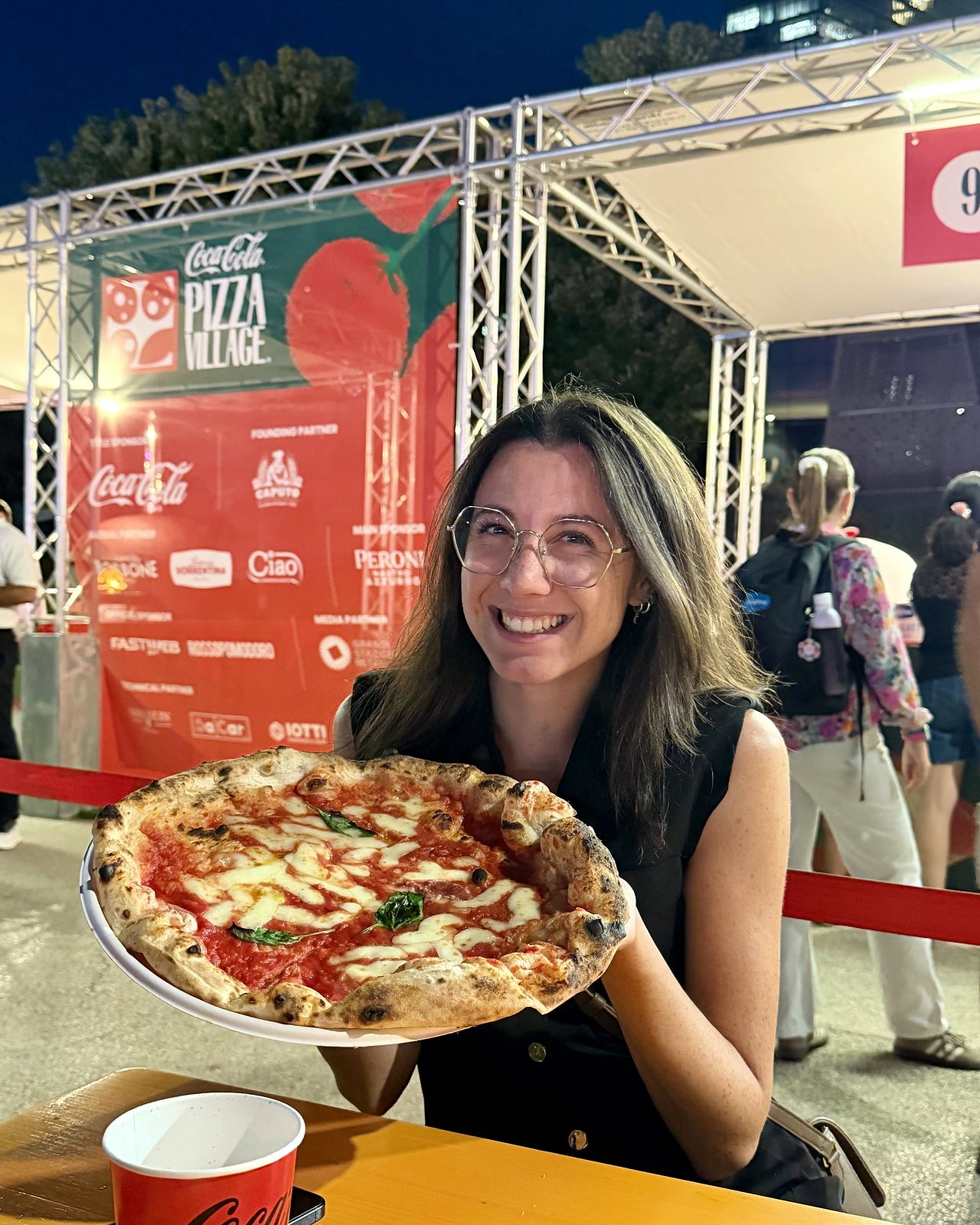 Ludovica con una pizza margherita in mano davanti alla scritta Coca-Cola Pizza Village a CityLife