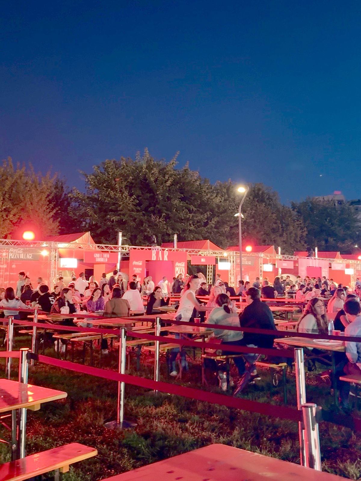 Il villaggio del Coca-Cola Pizza Village al Parco di CityLife a Milano all'imbrunire, con i tavoloni di legno e gli stand illuminati di rosso