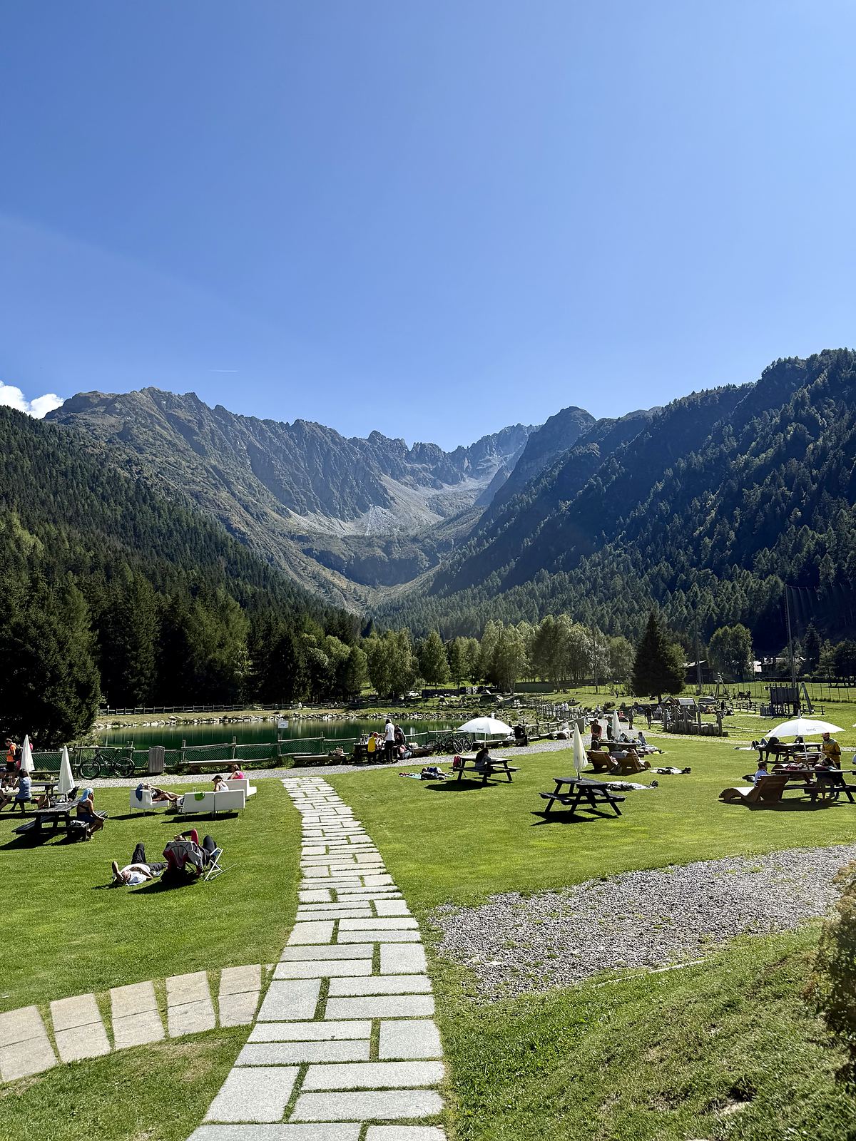 Prato di Valbione d'estate con lettini e tavoli da picnic davanti al lago e alle montagne dell'Adamello