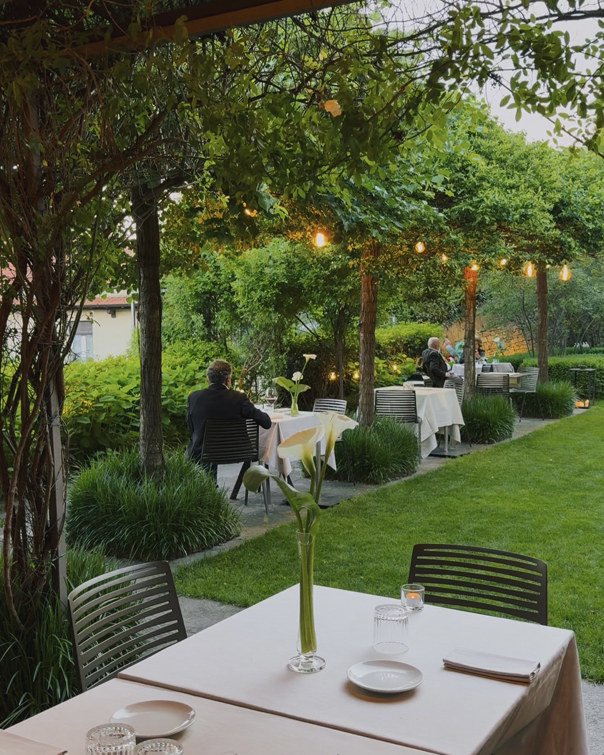 Giardino del Relais San Vigilio a Bergamo al tramonto, con tavoli apparecchiati, lucine e ospiti a cena nel verde